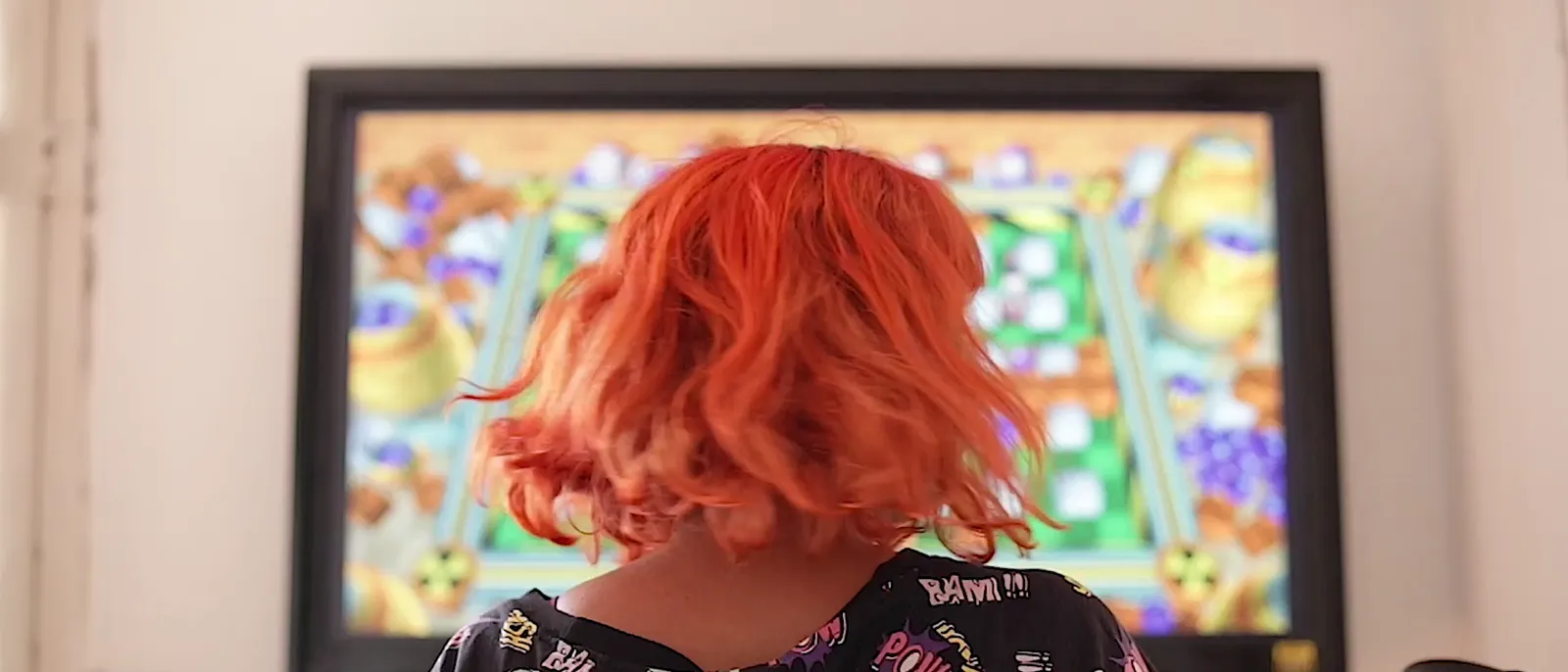 A person with bright orange hair, seen from behind, frustrated in front of a colourful video game on a TV